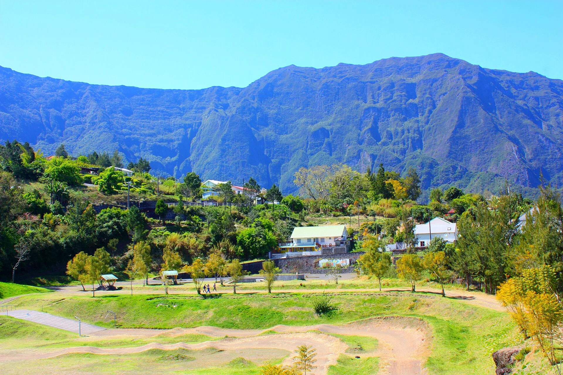 Parc verdoyant avec piste en terre et maisons au pied de hautes parois montagneuses sous un ciel bleu